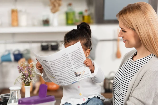 Joyeuse mère regardant adopté afro-américain gamin avec journal — Photo de stock