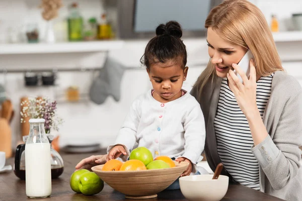 Glückliche Mutter spricht in der Nähe von adoptiertem afrikanisch-amerikanischem Kind in der Küche auf dem Smartphone — Stockfoto