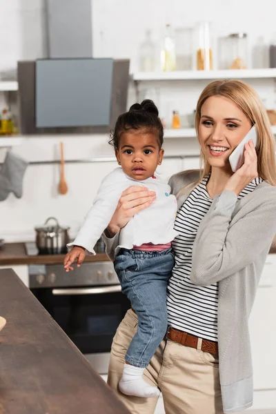 Счастливая мать разговаривает на смартфоне и держит усыновленного африканского американского ребенка на кухне — стоковое фото