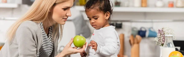 Madre che dona mela al bambino afroamericano adottato in cucina, banner — Foto stock