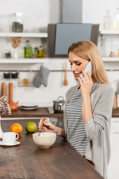 Femme blonde parlant sur téléphone portable et tenant cuillère près du bol — Photo de stock