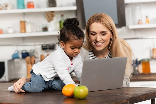 Freelancer feliz olhando para laptop perto da filha americana africana adotada na cozinha — Fotografia de Stock