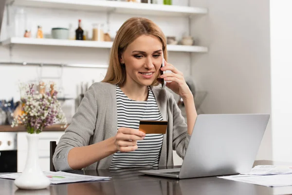 Happy woman holding credit card and talking on cellphone while shopping online — Stock Photo