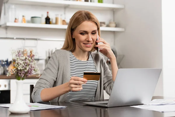Mujer feliz sosteniendo la tarjeta de crédito y hablando en el teléfono celular mientras que hace compras en línea - foto de stock