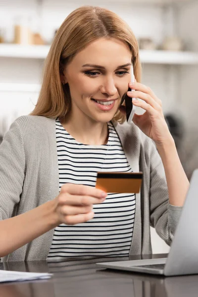 Mujer alegre sosteniendo la tarjeta de crédito y hablando en el teléfono celular mientras que hace compras en línea - foto de stock