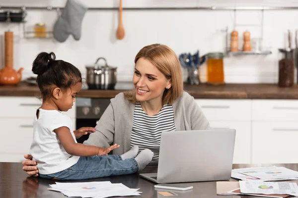 Smiling mother looking at adopted african american girl on table near gadgets — Stock Photo