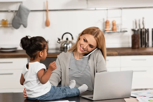 Sonriente madre hablando en el teléfono inteligente mirando adoptada chica afroamericana en la mesa cerca de la computadora portátil - foto de stock