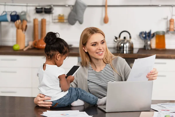 Mulher feliz olhando para o papel enquanto trabalhava em casa perto da filha americana africana adotada com smartphone — Fotografia de Stock