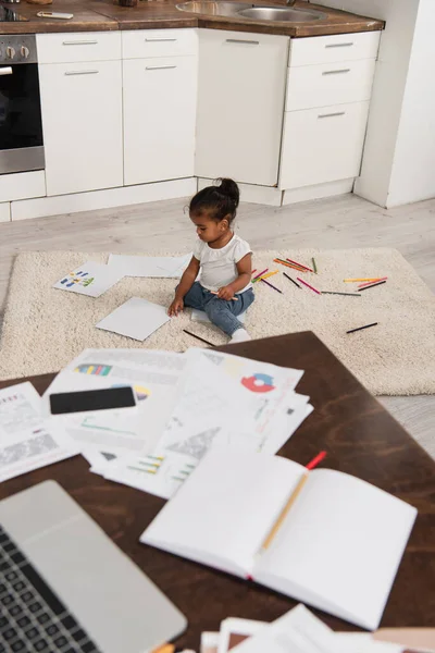 Menina criança afro-americana sentada no tapete perto de lápis coloridos e desenhos com gadgets borrados na mesa — Fotografia de Stock