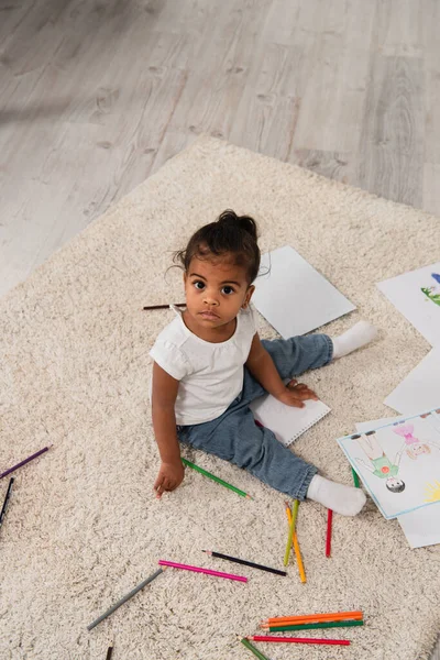 High angle view of african american toddler girl sitting on carpet near colorful pencils and drawings — Stock Photo