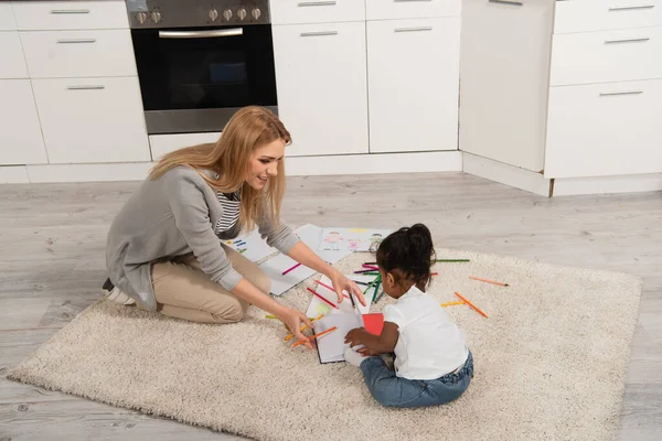 Joyeuse mère dessin avec adopté afro-américaine fille tout en étant assis sur le tapis — Photo de stock