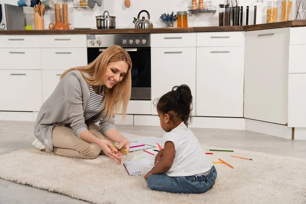 Happy mother drawing with adopted african american girl while sitting on carpet — Stock Photo