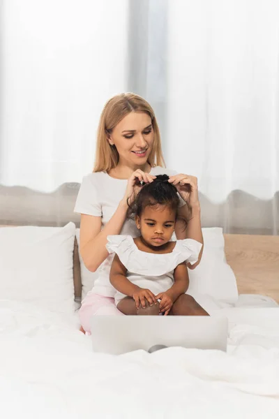 Heureux mère attacher les cheveux de adopté afro-américain enfant regarder film sur ordinateur portable dans la chambre — Photo de stock