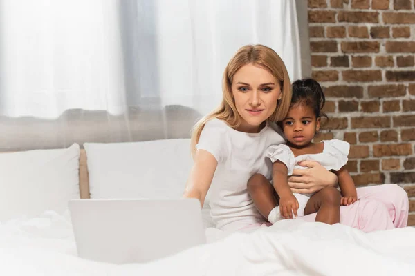 Cheerful mother and adopted african american watching film on laptop in bedroom — Stock Photo