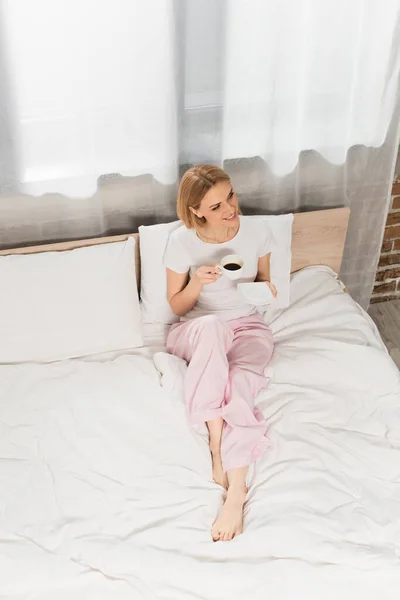 Vista de ángulo alto de la mujer rubia sonriente sosteniendo la taza de café en el dormitorio - foto de stock