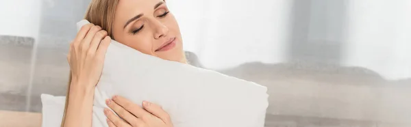 Happy blonde woman with closed eyes holding pillow in bedroom, banner — Stock Photo