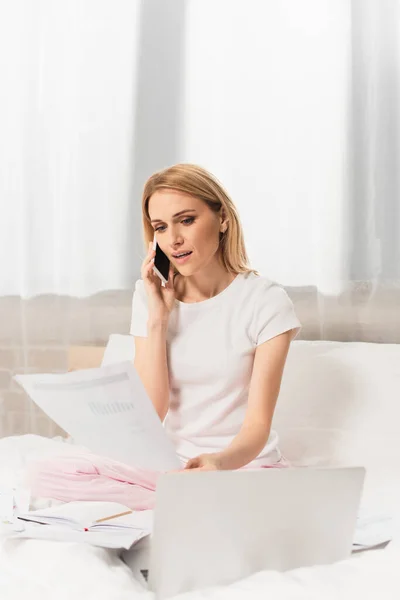 Блондинка розмовляє на мобільному телефоні біля ноутбука під час роботи з дому в спальні — стокове фото
