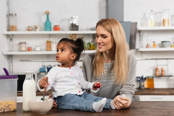 Niño afroamericano adoptado sosteniendo cuchara cerca de cuenco mientras está sentado en la mesa de la cocina cerca de madre feliz - foto de stock
