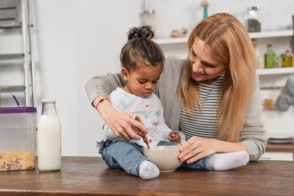 Adopted african american kid holding spoon near bowl while sitting on kitchen table near smiling mother — Stock Photo