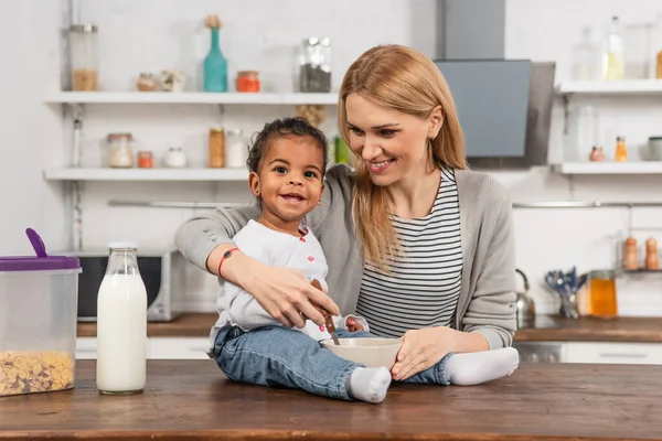 Felice adottato africano americano bambino tenendo cucchiaio vicino ciotola mentre seduto sul tavolo della cucina vicino sorridente madre — Foto stock