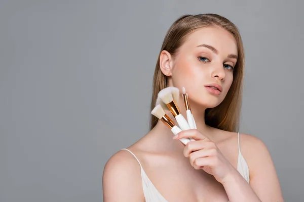 Pretty young woman holding set of different cosmetic brushes isolated on grey — Stock Photo