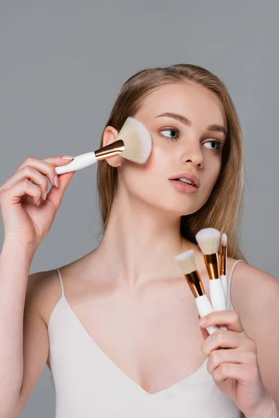 Mujer joven sosteniendo conjunto de diferentes cepillos cosméticos y aplicando polvo facial aislado en gris - foto de stock