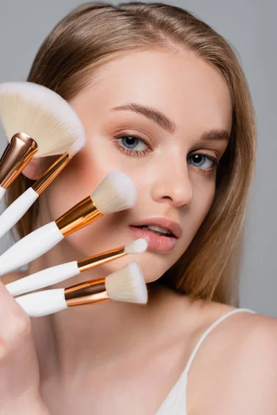 Mujer joven sosteniendo conjunto de diferentes cepillos cosméticos mientras mira la cámara aislada en gris - foto de stock