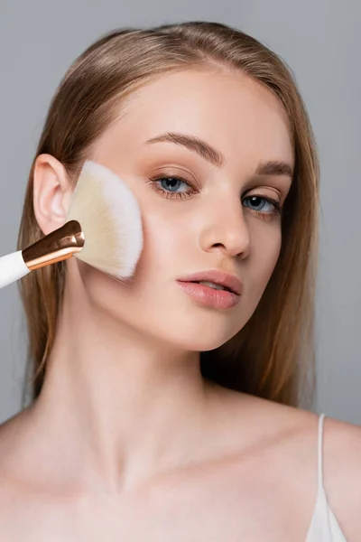 Mujer joven sosteniendo cepillo cosmético y aplicando polvo facial aislado en gris - foto de stock