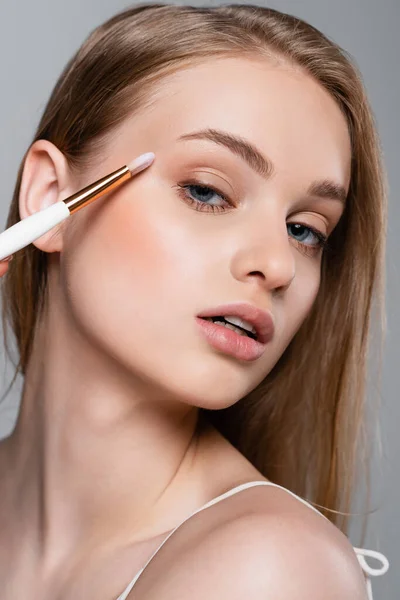 Primer plano de mujer joven sosteniendo cepillo cosmético y aplicando sombra de ojos aislado en gris - foto de stock