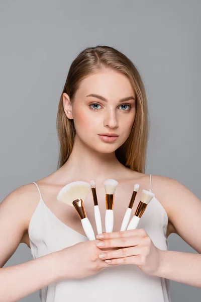 Mujer joven sosteniendo diferentes cepillos cosméticos aislados en gris - foto de stock