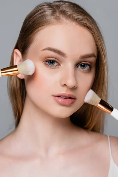 Close up of young woman holding different cosmetic brushes and applying face powder isolated on grey — Stock Photo
