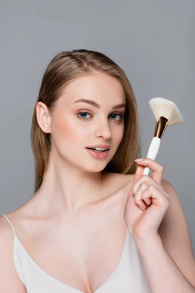 Happy young woman holding cosmetic brush isolated on grey — Stock Photo