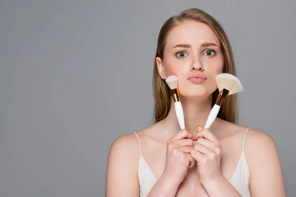 Jeune femme tenant des pinceaux cosmétiques près du visage isolé sur gris — Photo de stock