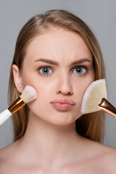 Young woman holding cosmetic brushes and applying face powder isolated on grey — Stock Photo