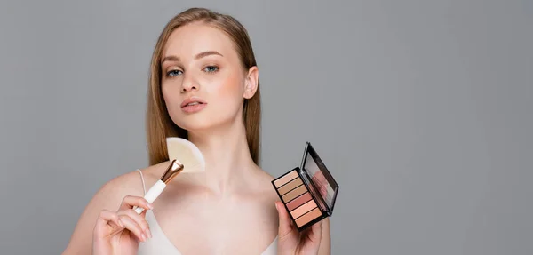 Young woman holding cosmetic brush and eye shadow and blush palette isolated on grey, banner — Stock Photo