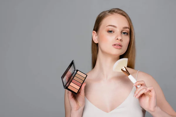 Pretty woman holding cosmetic brush and eye shadow and blush palette isolated on grey — Stock Photo