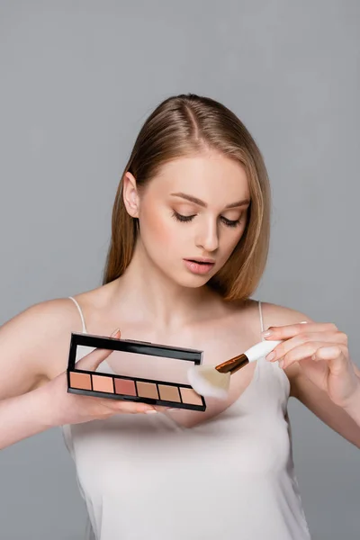 Jeune femme tenant pinceau cosmétique appliquant le fard à joues de la palette isolé sur gris — Photo de stock