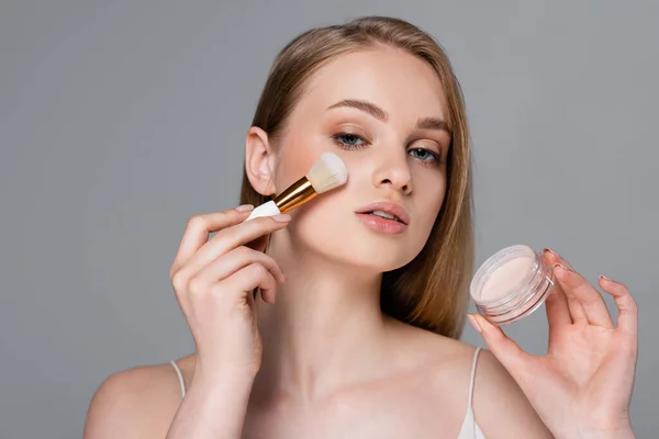 Mujer joven sosteniendo cepillo cosmético mientras aplica polvo facial y contenedor aislado en gris - foto de stock