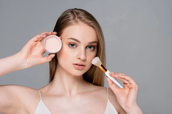 Junge Frau hält Kosmetikpinsel und Gesichtspuder isoliert auf grau — Stockfoto