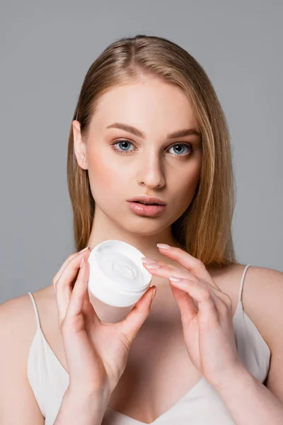 Pretty young woman holding container with cosmetic cream isolated on gray — Stock Photo