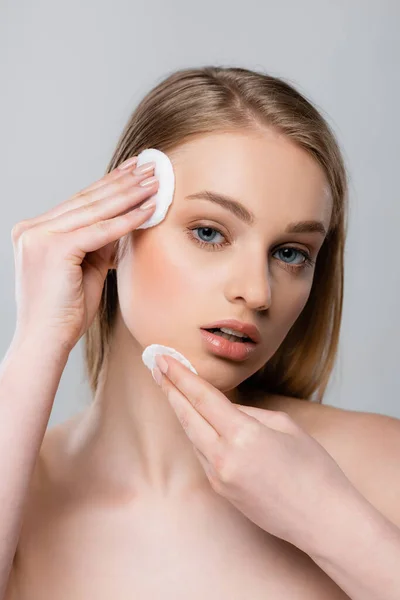 Mujer joven con hombros desnudos quitando el maquillaje con almohadillas de algodón aisladas en gris - foto de stock