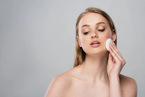 Young woman with bare shoulders removing makeup with cotton pad isolated on gray — Stock Photo