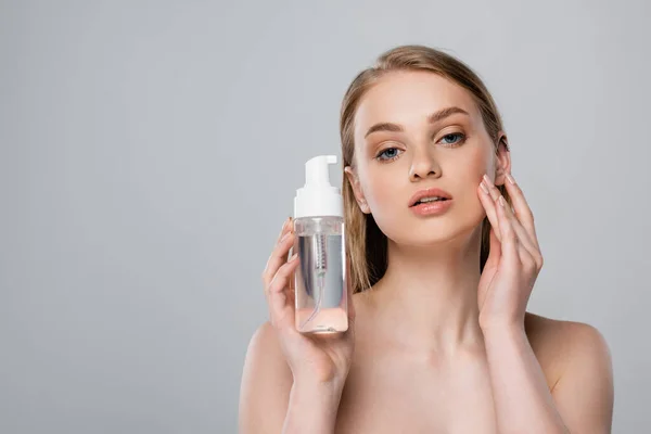 Pretty young woman with bare shoulders holding bottle with micellar water isolated on grey — Stock Photo