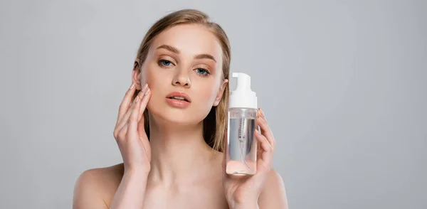 Pretty young woman with bare shoulders holding bottle with micellar water isolated on grey, banner — Stock Photo
