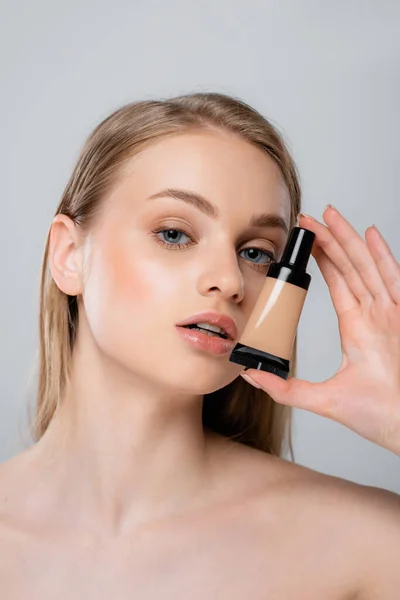 Pretty woman with bare shoulders holding tube with makeup foundation isolated on grey — Stock Photo