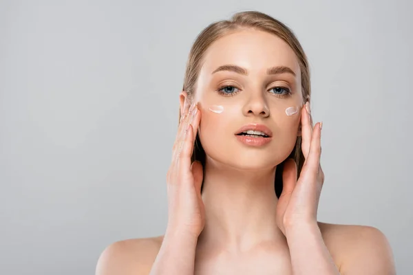 Jeune femme aux yeux bleus appliquant de la crème cosmétique sur le visage isolé sur gris — Photo de stock