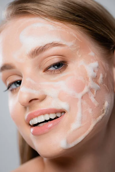 Close up of smiling woman with blue eyes and cleansing foam on face isolated on grey — Stock Photo
