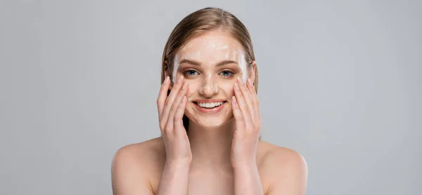 Positive junge Frau mit blauen Augen, die Reinigungsschaum auf das Gesicht aufträgt, isoliert auf grau, Banner — Stockfoto