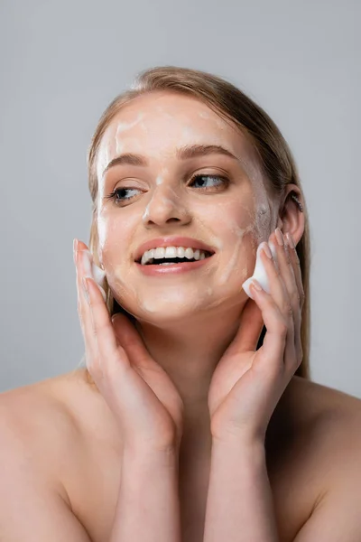 Joyeuse jeune femme aux yeux bleus et mousse nettoyante sur le visage isolé sur gris — Photo de stock