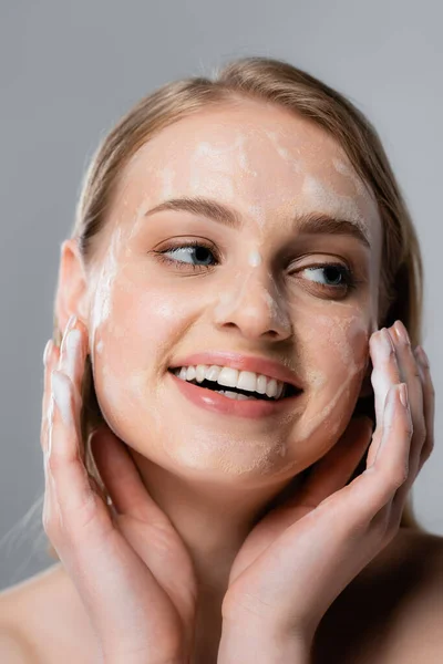 Giovane donna positiva con occhi azzurri e schiuma detergente sul viso isolata sul grigio — Foto stock
